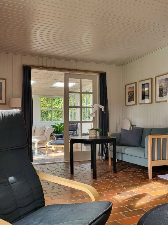 Blå sofa med kig til den lyse tilbygning, Langelandsfotos på væggen og blomst på sofabordet i sommerhuset på Langeland.