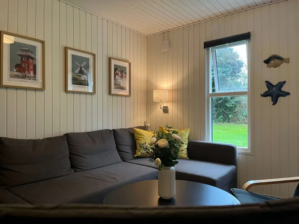 Sofa med gule puder, blomster og Langelandsfoto på væggen i hyggelig stue i sommerhuset på Langeland.